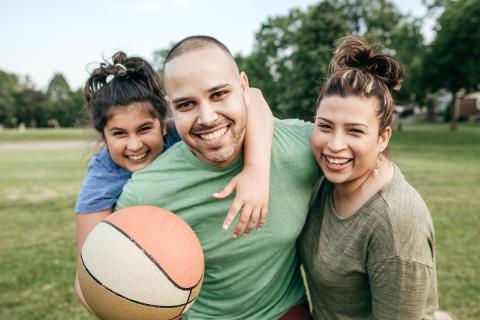 A smiling dad, mom, and female child play basketball in a public park, enjoying physical activity together
