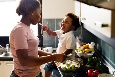 A mother prepares a healthy green salad while her toddler snacks on fruit on the kitchen counter