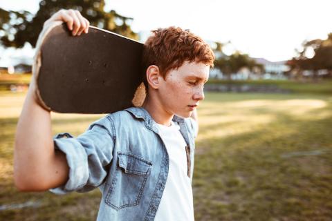 Adolescent boy with skateboard in rural area, with his eyes closed, looking sad.