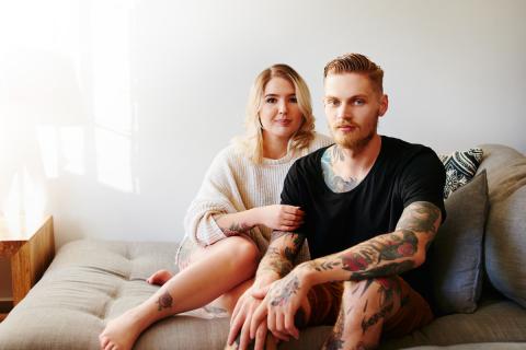 A young couple sits on a chaise lounge staring intently at the camera and not communicating